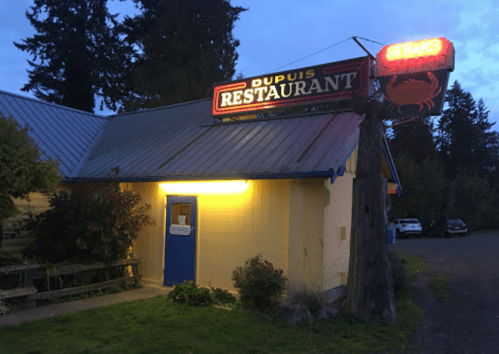A cozy restaurant with a neon sign reading "Dupuis Restaurant" and "Steaks & Seafood," surrounded by trees at dusk.