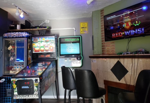 A cozy game room featuring a pinball machine, arcade game, and a TV displaying a game score with "RED WINS!"