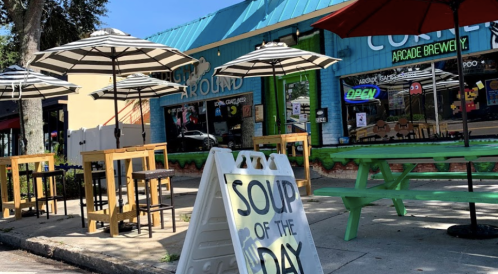 Outdoor seating area with umbrellas in front of a colorful arcade brewery, featuring a sign for "Soup of the Day."
