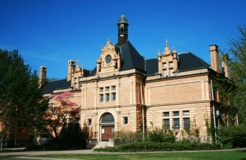 Historic brick building with ornate architecture, surrounded by greenery and blue sky.