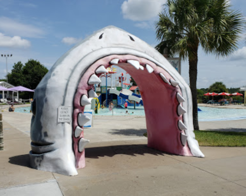 A large shark mouth entrance at a water park, with a pool and palm trees in the background.