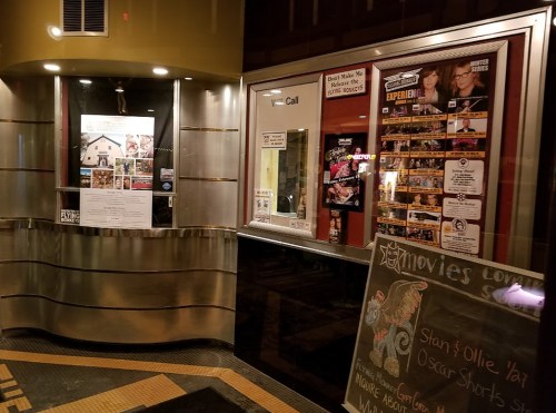 A movie theater entrance with posters, a chalkboard sign, and a ticket booth displaying upcoming films and events.