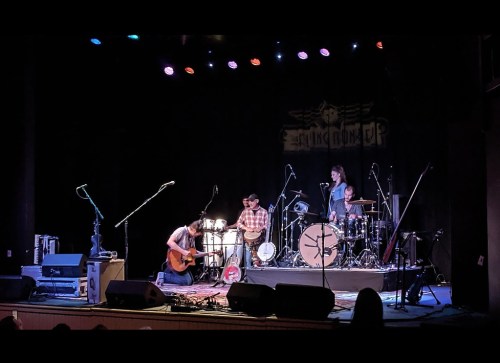 A band performs on stage with a guitar, drums, and percussion instruments under colorful lights.