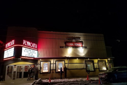 The exterior of a theater at night, featuring a marquee and bright lights, with people gathered outside.