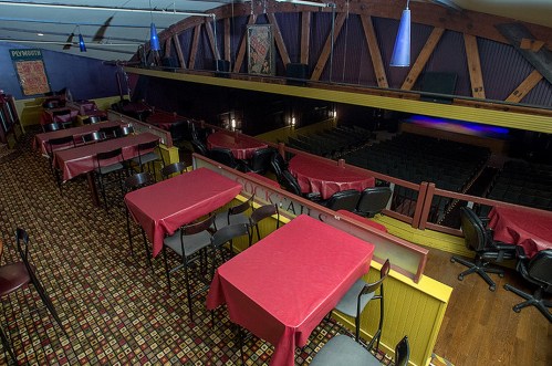 Interior of a theater with red tables and chairs, featuring a stage area and wooden beams overhead.