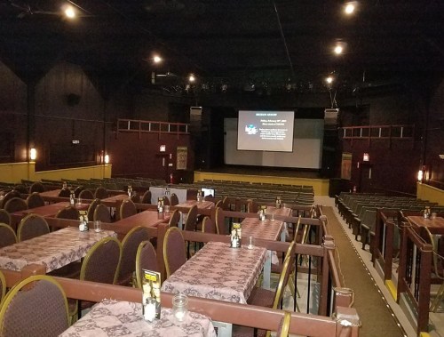 Interior of a theater with empty tables and chairs, a stage in the background displaying a presentation screen.