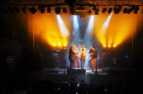 A live music performance on stage with three musicians and vibrant yellow lighting, while an audience watches below.