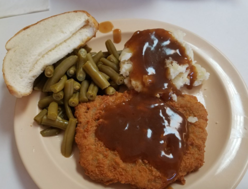 A plate with fried chicken covered in gravy, mashed potatoes, green beans, and a slice of bread.
