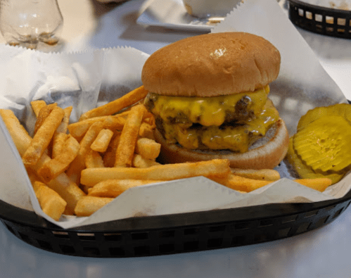 A cheeseburger with two patties and melted cheese, served with a side of golden fries and a pickle slice in a basket.