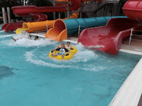 Two children ride a yellow inner tube down a water slide, splashing in a pool with colorful slides in the background.