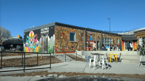 Colorful mural on a building exterior with outdoor seating, showcasing vibrant art and a lively atmosphere.