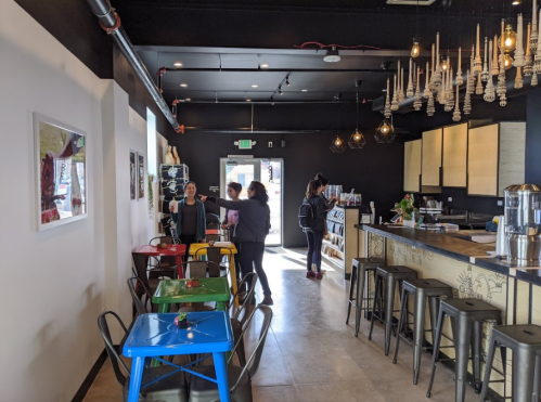 A modern café interior with colorful tables, a counter, and customers interacting. Bright lighting and decor enhance the ambiance.