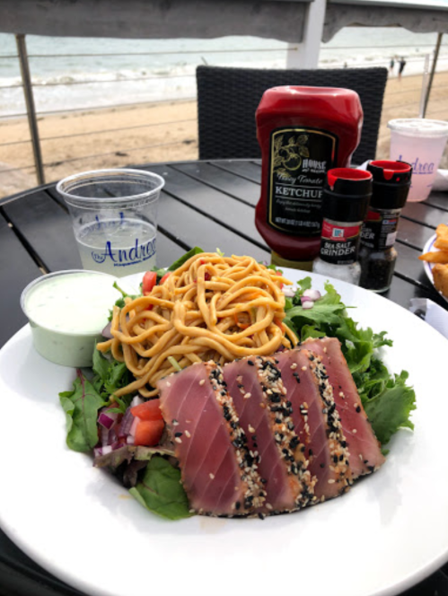 A plate with seared tuna, salad, noodles, and condiments, set against a beach view.