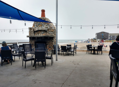 Outdoor seating area with a stone fireplace, overlooking the beach, under blue canopies and string lights.
