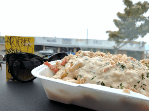 A close-up of loaded fries topped with creamy sauce and herbs, with sunglasses and a business card in the background.