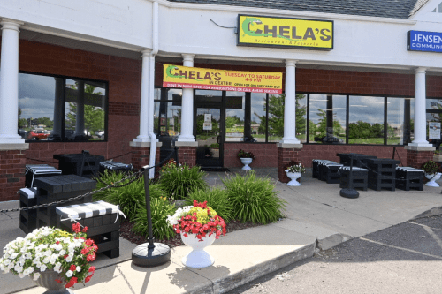 Exterior of Chela's Restaurant with outdoor seating, planters, and a sign displaying hours and contact information.