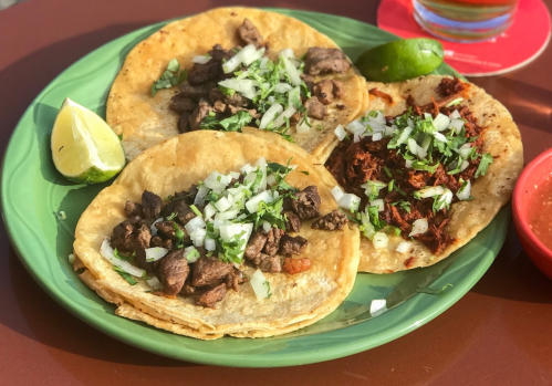 Three tacos on a green plate: one with beef, one with shredded meat, topped with onions and cilantro, with lime and salsa.