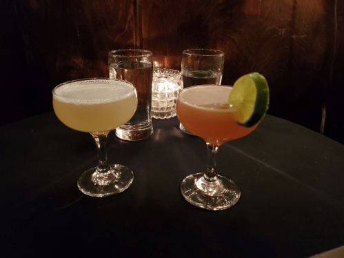 Two cocktails on a black table, one light yellow and one pink with a lime wedge, alongside glasses of water and a candle.