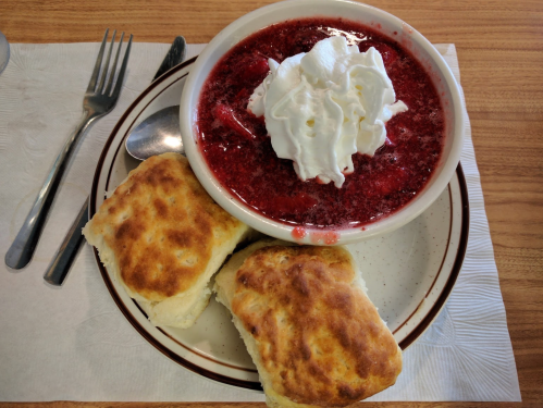 A bowl of strawberry dessert topped with whipped cream, served with two golden biscuits on a plate.