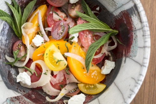 A colorful salad with various tomatoes, onions, mint leaves, and crumbled cheese in a decorative bowl.