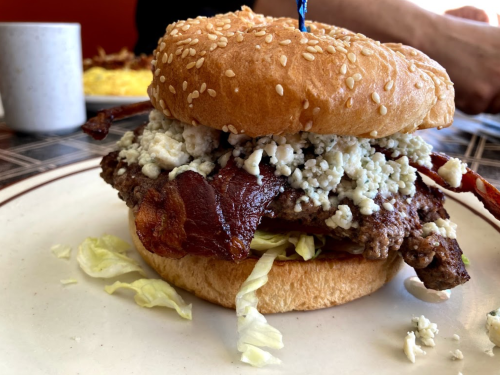 A sesame seed bun holds a bacon cheeseburger topped with blue cheese and lettuce, served on a plate.