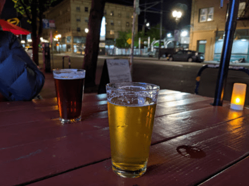 Two glasses of beer on a wooden table, with a city street and buildings softly lit in the background at night.