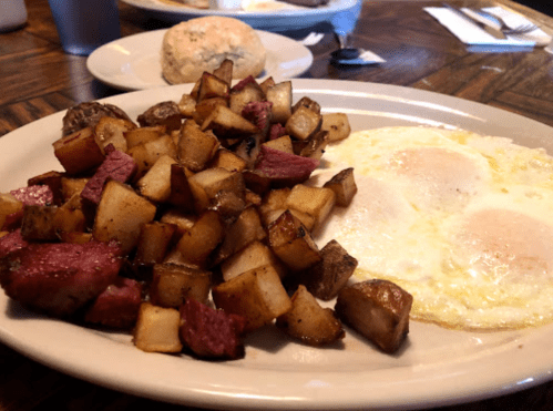 A plate of breakfast featuring two fried eggs, diced potatoes, and chunks of meat, with a biscuit in the background.