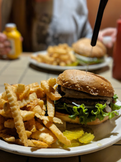 A close-up of a sesame seed burger with lettuce and pickles, served with crinkle-cut fries on a plate.