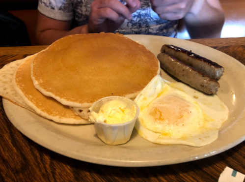 A plate with three pancakes, two sausage links, and a fried egg with a small cup of butter.