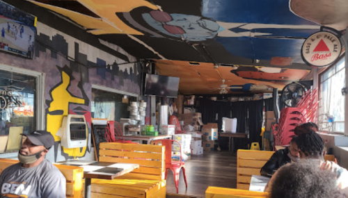 Interior of a colorful restaurant with mural art, wooden tables, and chairs, featuring a TV and stacked boxes in the background.