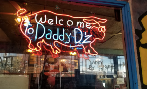 Neon sign of a cow with the text "Welcome to Daddy D'z" in a barbecue restaurant window.