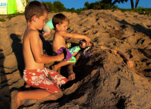 Two young boys play in the sand, burying a third child, with a sunny beach scene in the background.