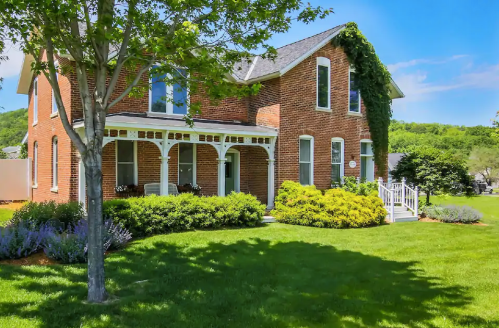 A charming brick house with a porch, surrounded by lush greenery and colorful flowers under a clear blue sky.