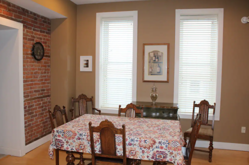 A dining room with a patterned tablecloth, wooden chairs, brick wall, and two windows with blinds.