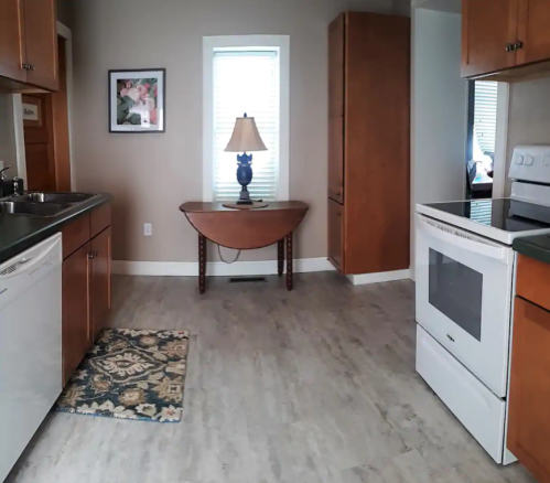 A bright kitchen with wooden cabinets, a table, a lamp, and a rug, featuring a stove and dishwasher.