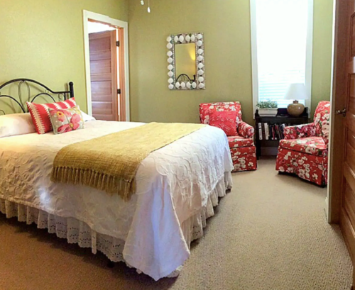 Cozy bedroom with a bed, floral chairs, and a mirror, featuring soft green walls and warm lighting.