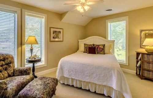 Cozy bedroom with a white bed, floral accents, armchair, and natural light from windows.
