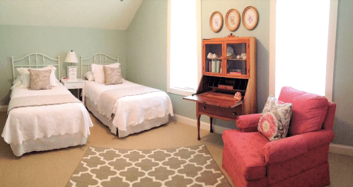 A cozy bedroom with two twin beds, a wooden desk, a pink armchair, and decorative wall art.