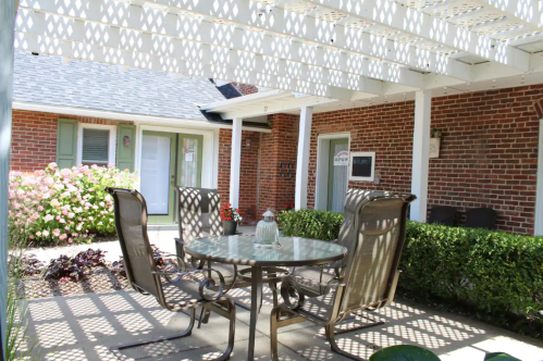A patio with a glass table and chairs, shaded by a lattice, surrounded by greenery and blooming flowers.