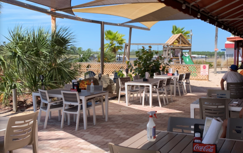 Outdoor dining area with tables, palm trees, and a playground in the background under a shaded canopy.