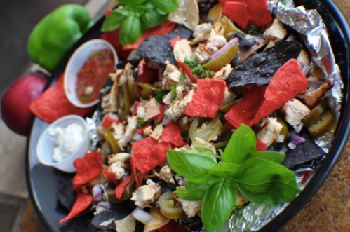 A colorful platter of nachos topped with chicken, jalapeños, red and blue tortilla chips, and fresh basil.