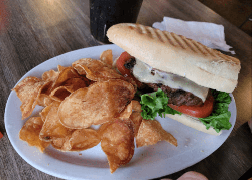 A grilled sandwich with lettuce, tomato, and cheese, served with a side of crispy potato chips and a drink.