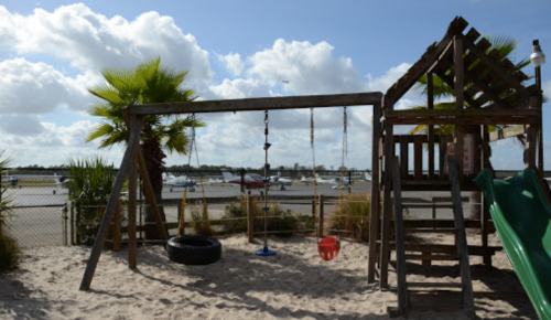 Playground with swings and a slide, surrounded by palm trees, overlooking a runway with parked planes in the background.