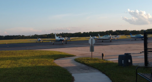 Several small airplanes parked on a runway with a grassy area and trees in the background under a clear sky.