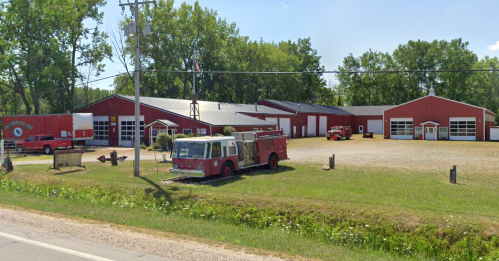 A red fire station with vintage fire trucks parked outside, surrounded by trees and a gravel lot.