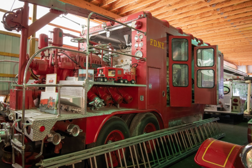 A vintage red fire truck inside a barn, featuring various hoses and equipment, with a toy fire truck on display.