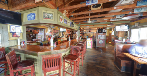 A cozy restaurant interior featuring a wooden bar, high chairs, and rustic decor with various signs and TVs.