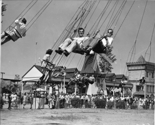 Black and white photo of people on a swing ride at a fair, with a crowd and amusement park structures in the background.