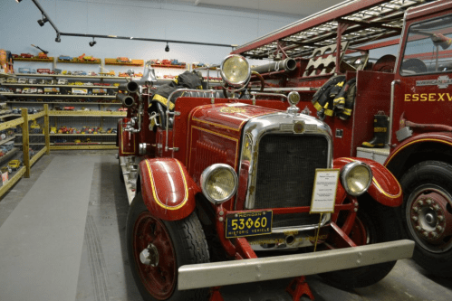 A vintage red fire truck displayed in a museum, with firefighting gear and toy vehicles in the background.