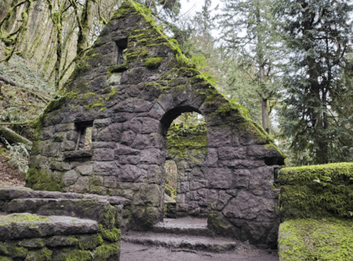 Ruins of a moss-covered stone structure surrounded by lush greenery and trees in a forested area.